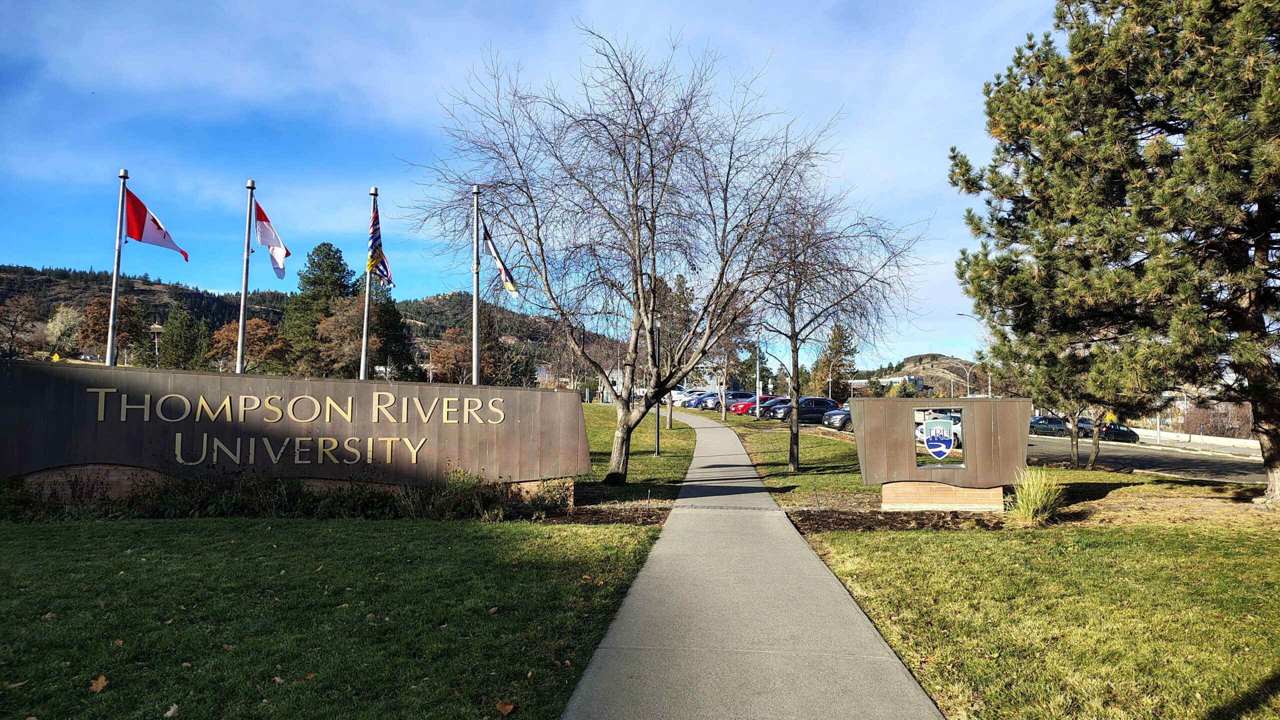Aerial view of Kamloops, British Columbia, showing the Thompson River winding through the city centre, with the surrounding hills and mountains of the Interior in the background. The research site, Thompson Rivers University, sits on the hillside on the right.