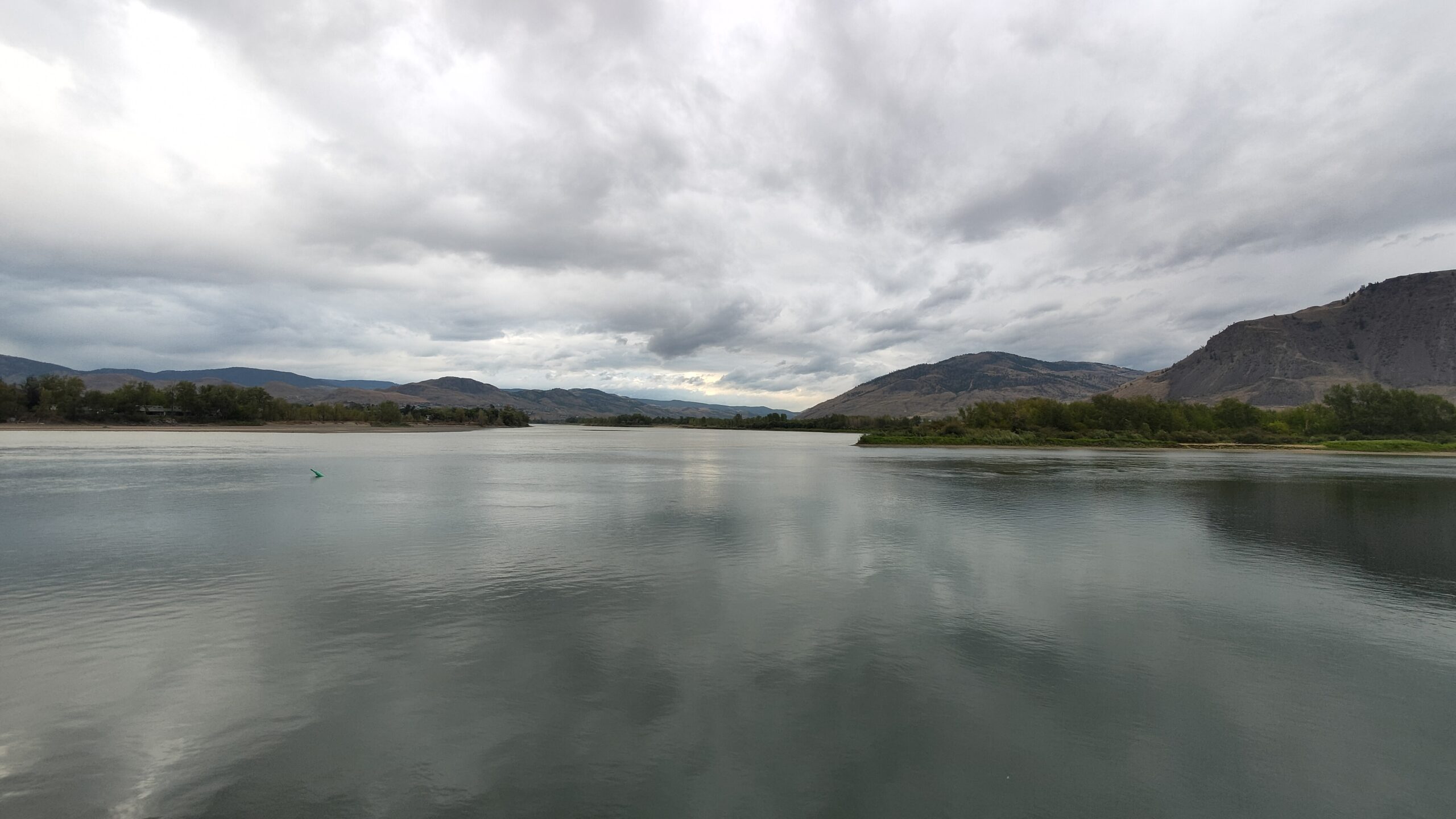The South Thompson River at Kamloops framed by the silhouetted branches of cottonwood trees in the foreground. A dramatic storm-heavy sky fills most of the frame. A metal dock railing is visible at the water's edge. The river stretches back toward rolling hills.
