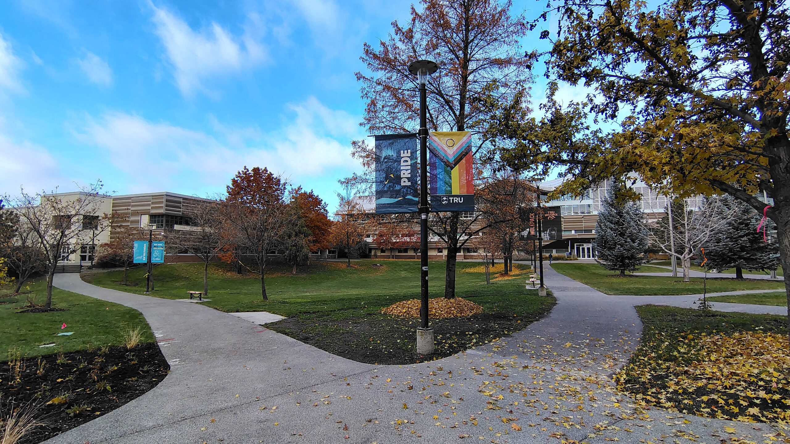 A lamppost at Thompson Rivers University displays a large Pride banner with rainbow colours and the TRU logo. Autumn-coloured trees and empty campus pathways surround it.