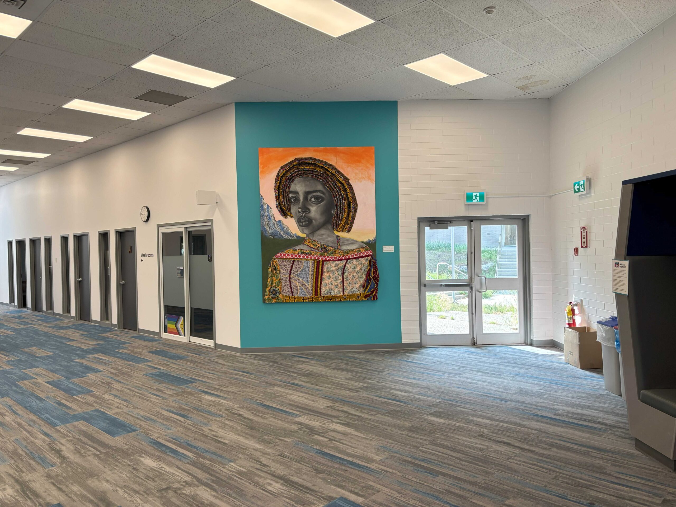 An empty institutional corridor inside a Thompson Rivers University building. A large, vibrant painting of a young Black woman wearing a golden halo-like crown of braids and traditional patterned dress hangs on a teal accent wall. The corridor is empty. Fluorescent lights illuminate the space. An ATM is partially visible at the far right edge of the frame.