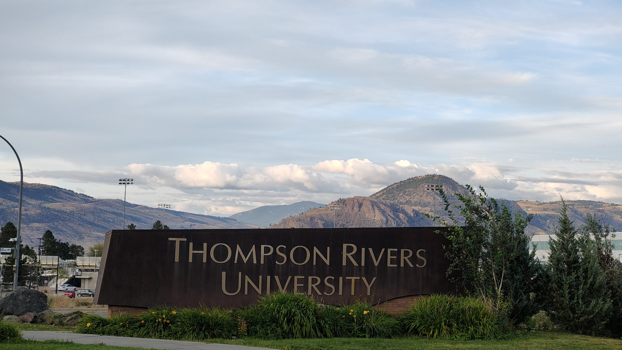 Thompson Rivers University entrance sign with Canadian, British Columbia, and other flags flying against a blue sky. A pathway leads into the campus grounds.
