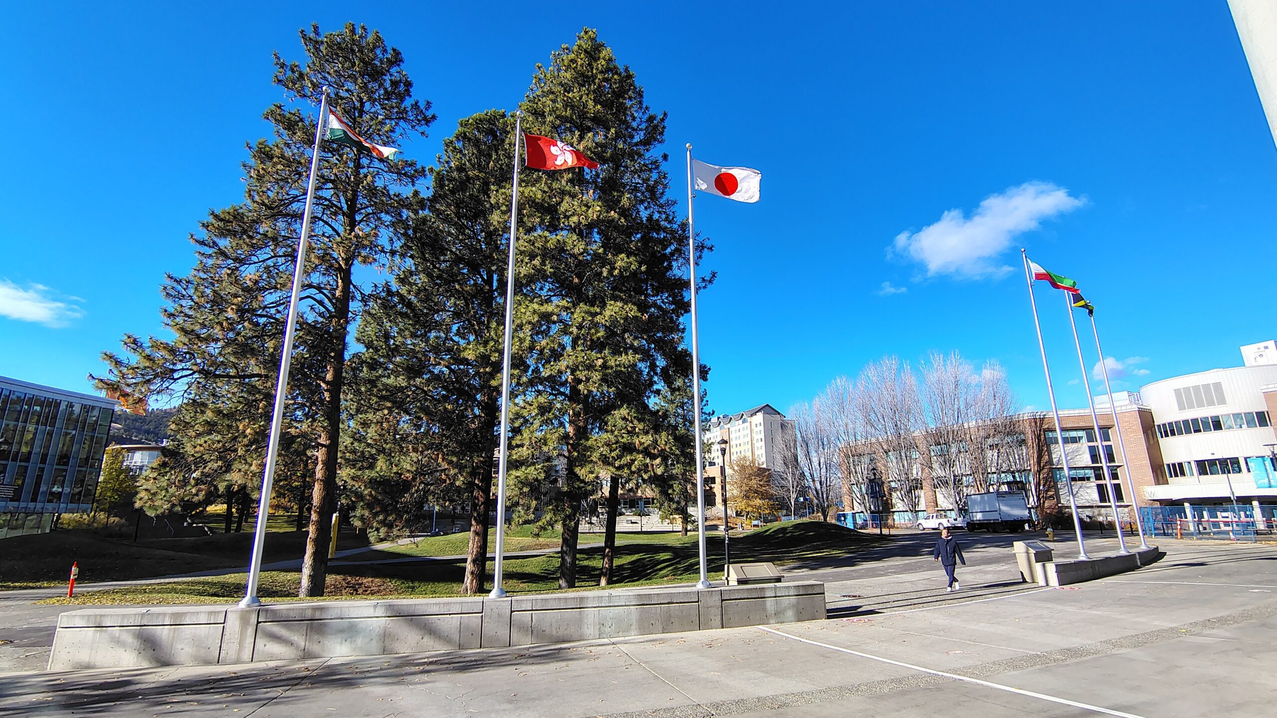 Five international flags flying on tall flagpoles at Thompson Rivers University. A single person in dark clothing walks alone across the wide empty plaza in the foreground. The flags of India, Hong Kong, Japan, and others are visible. Student residences appear in the background.