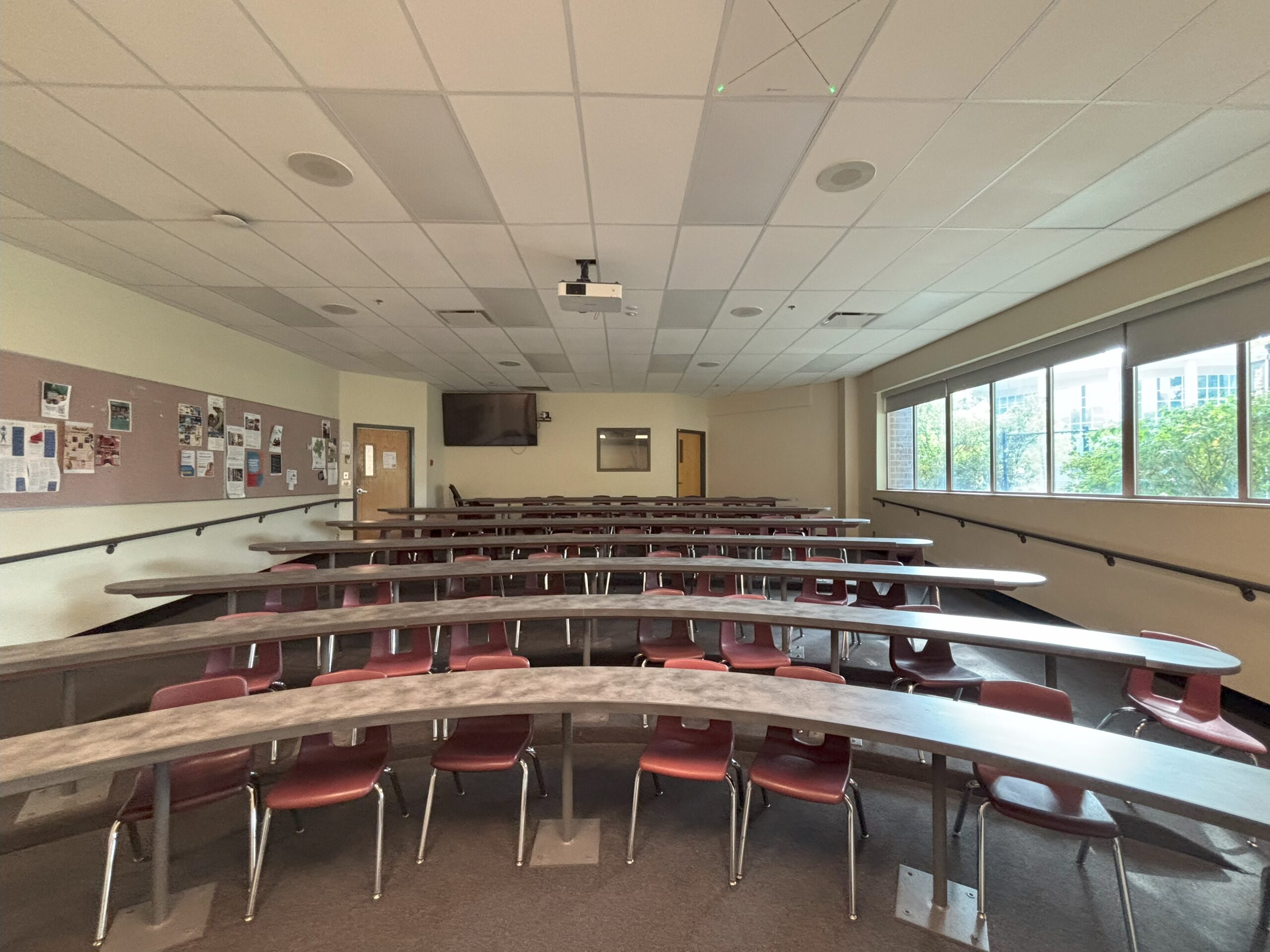 An empty, curved tiered lecture hall at Thompson Rivers University, with blue accent walls, curved rows of desks facing a whiteboard and projector screen at the front. Every seat is unoccupied.