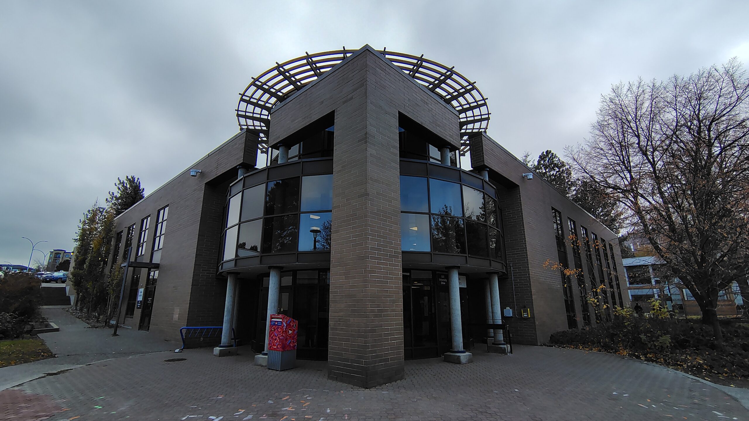 An empty tiered lecture hall at Thompson Rivers University with curved rows of red plastic chairs, a corkboard bulletin board covered in notices on the left wall, and large windows on the right letting in natural light.