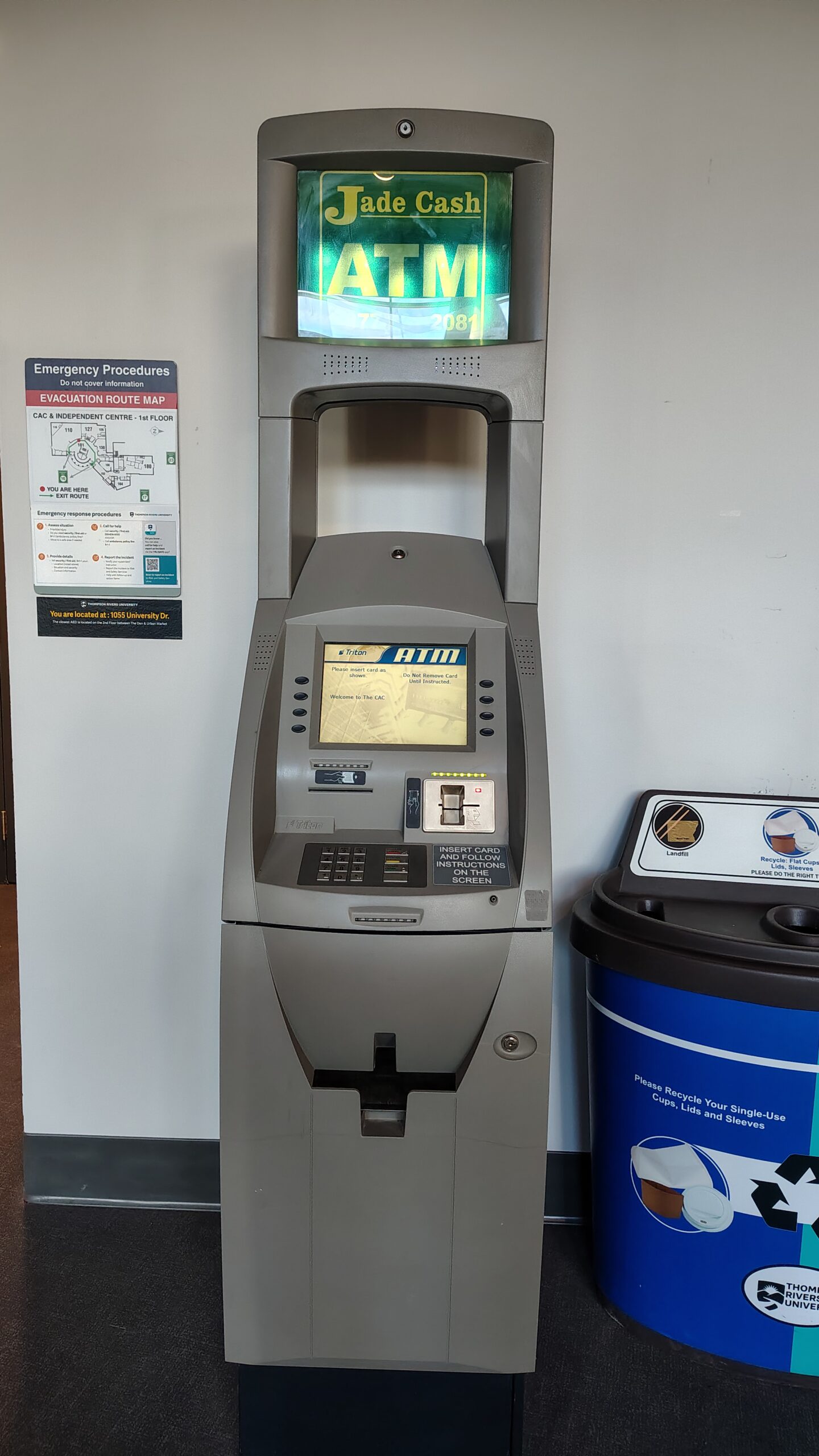 The Brown Family House of Learning at Thompson Rivers University: a striking glass curtain-wall building with a curved roofline, a silhouette of a wolf sculpture visible at the roofline, green lawns in the foreground, autumn trees to either side, and a bright blue sky above.