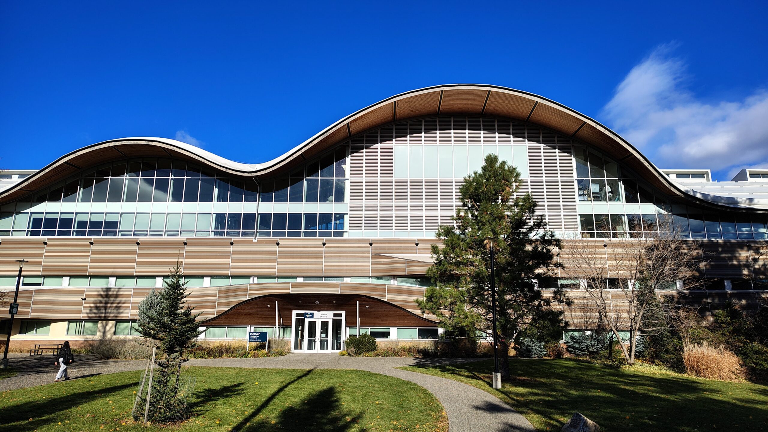 Thompson Rivers University's Old Main building, with its distinctive double-wave roofline of wood and glass, surrounded by manicured grounds and autumn-tinged trees under a bright blue sky.
