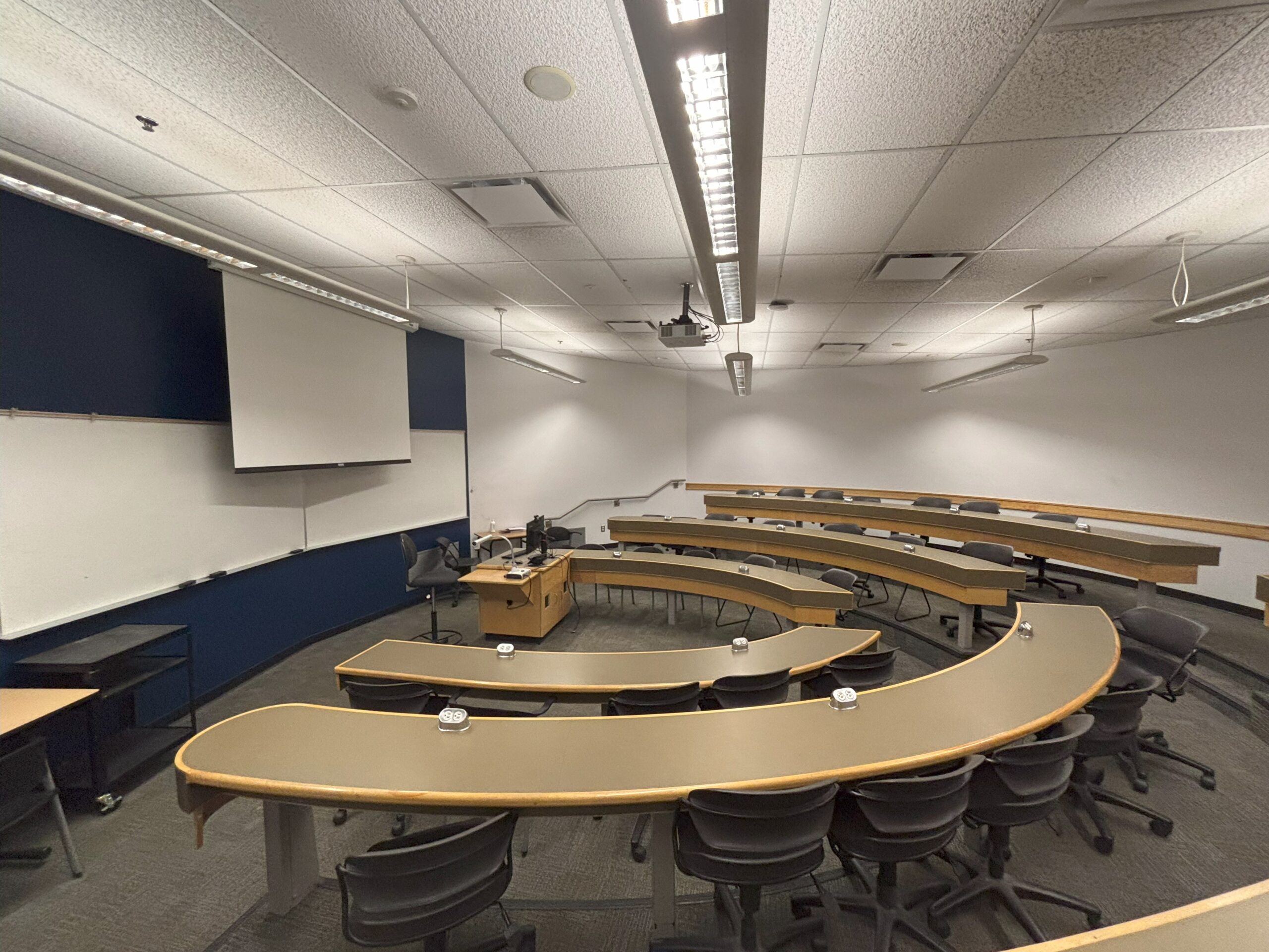 The interior of a student lounge at Thompson Rivers University: warm wood tables, teal-blue walls, large windows letting in daylight, and a handful of students seated at tables. The space is bright, inviting, and largely empty.